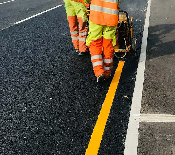 Two workers in reflective safety gear painting a bright yellow line on fresh asphalt using a line striping machine