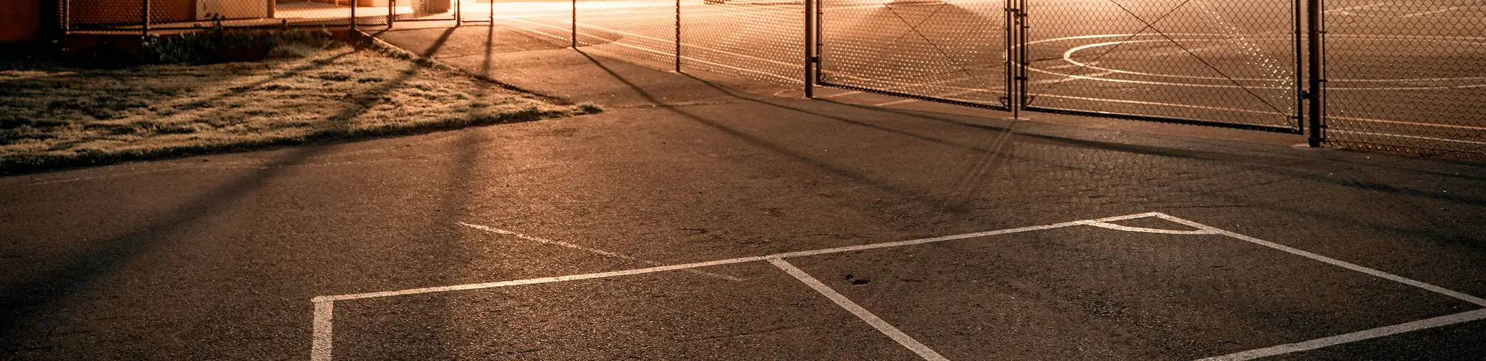 Freshly paved school basketball court with clean white lines under warm sunlight, enclosed by a tall chain-link fence, representing safe and durable surfaces for educational institutions.