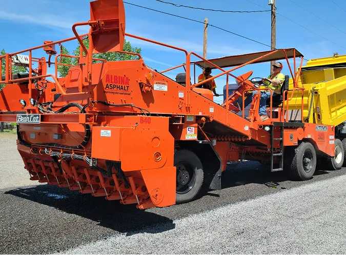 Bright orange asphalt paving machine operated by crew members laying new roadway surface under clear blue skies