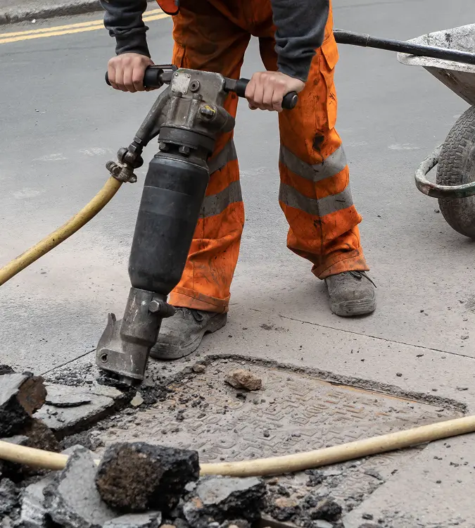 Worker using a jackhammer to break asphalt pavement during demolition service, with debris scattered on the road surface.