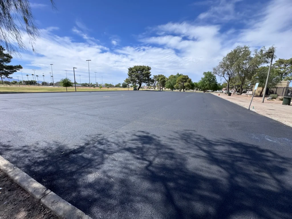 Freshly paved parking lot at El Prado Park with park trees and sports field lights in the background
