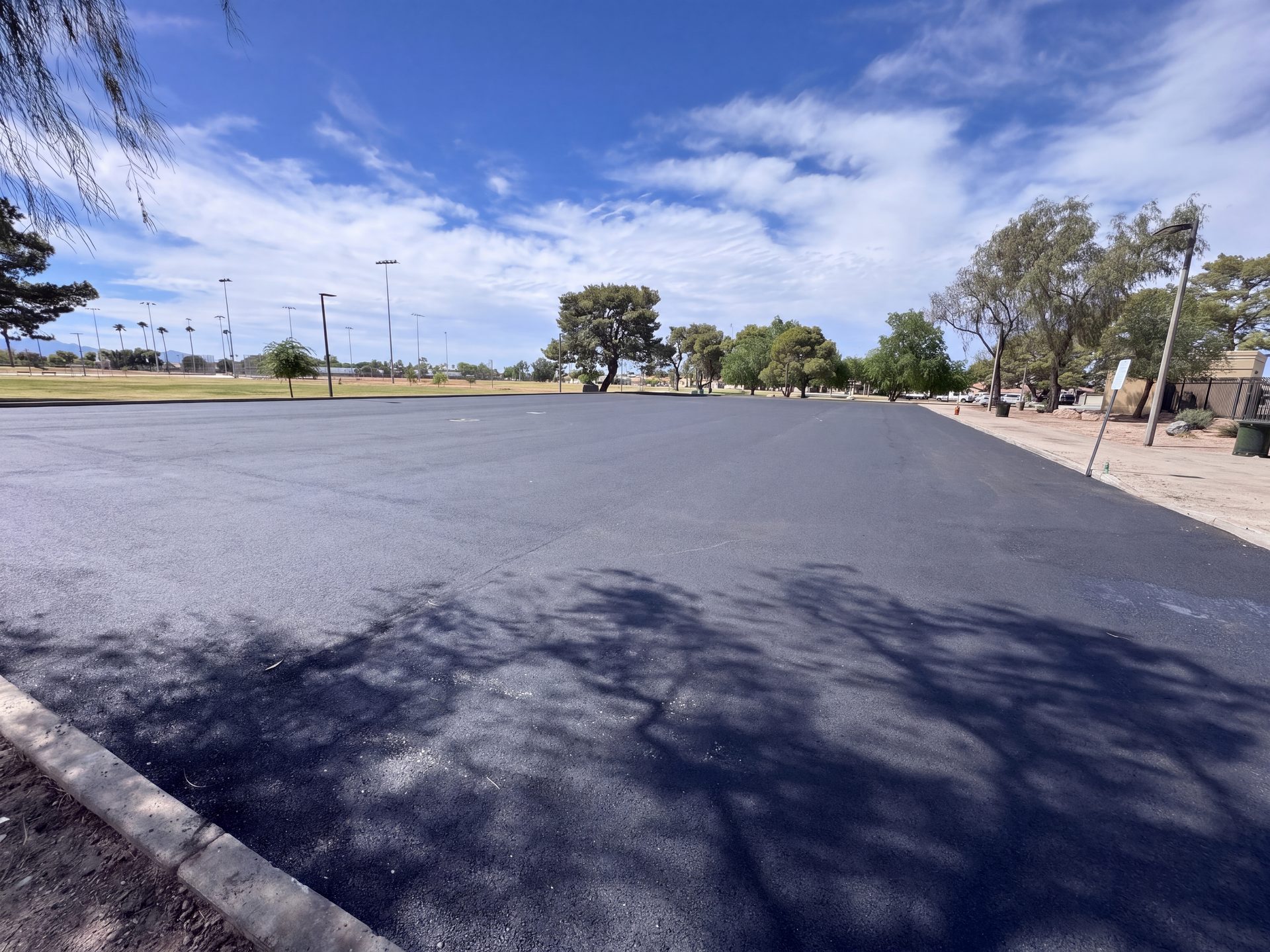 Freshly paved parking lot at El Prado Park with park trees and sports field lights in the background