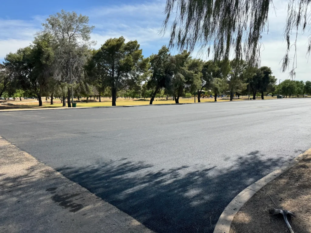 Wide view of El Prado Park parking lot during asphalt paving with mature trees lining the lot