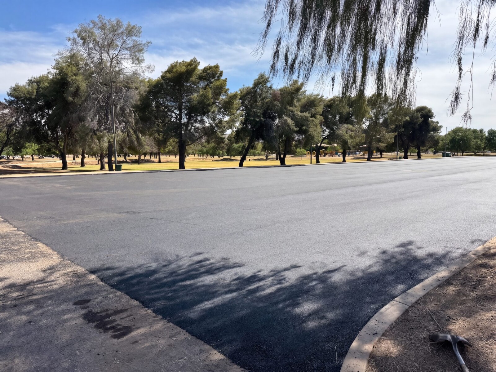 Wide view of El Prado Park parking lot during asphalt paving with mature trees lining the lot