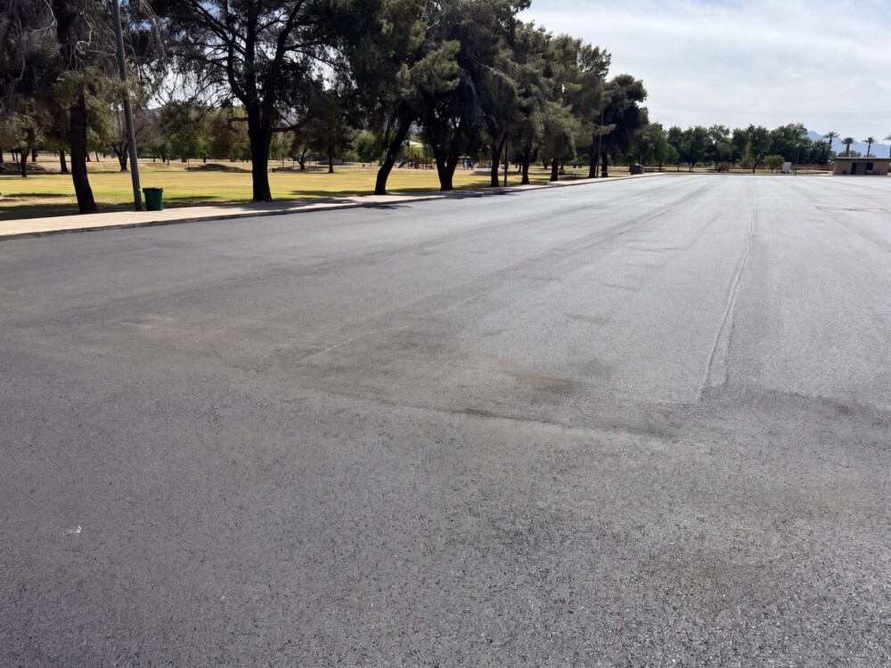 Wide view of El Prado Park parking lot during asphalt paving with mature trees lining the lot