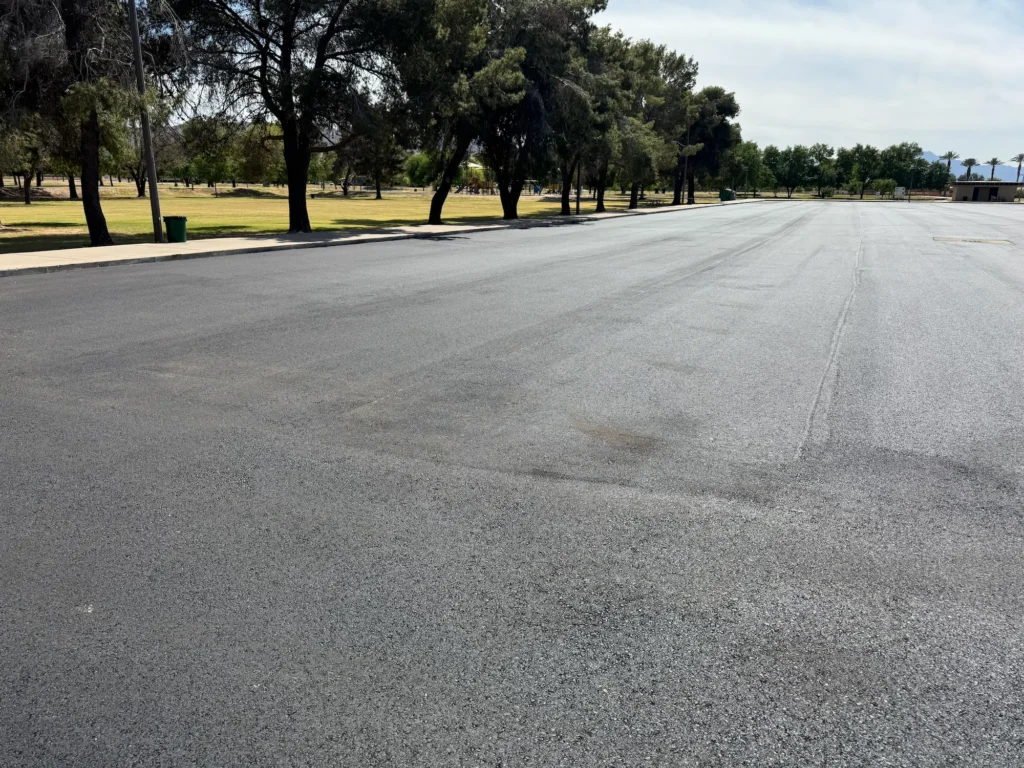 Wide view of El Prado Park parking lot during asphalt paving with mature trees lining the lot