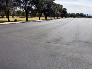 Wide view of El Prado Park parking lot during asphalt paving with mature trees lining the lot
