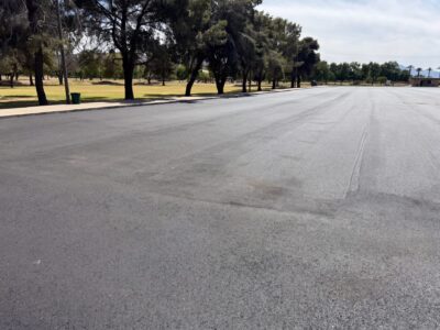 Wide view of El Prado Park parking lot during asphalt paving with mature trees lining the lot